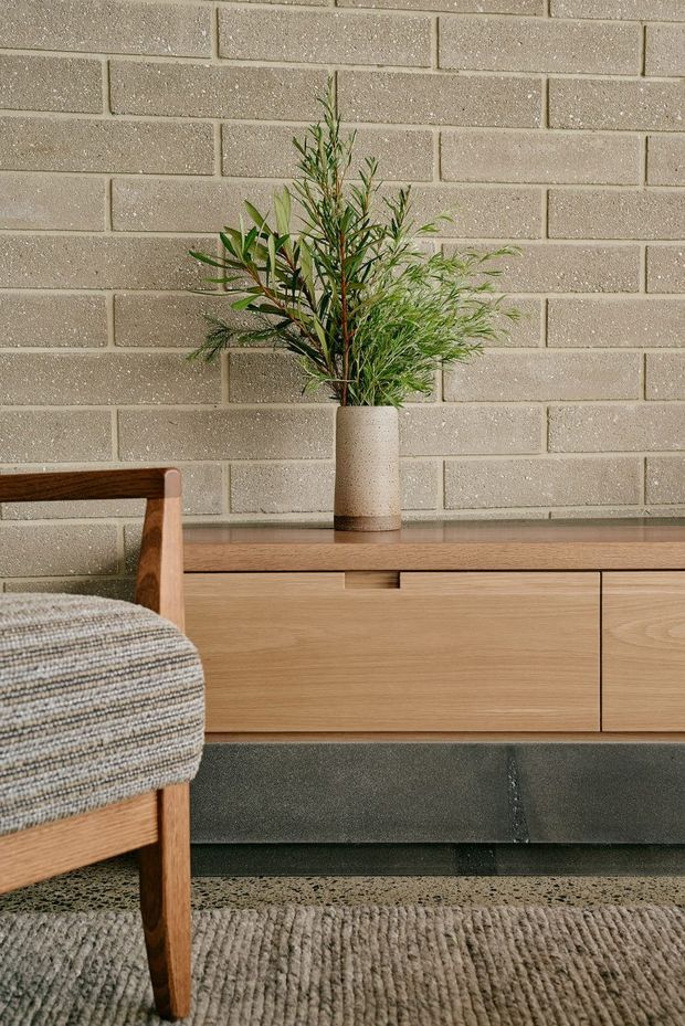 Close-up interior vignette featuring a mid-century-inspired timber armchair, a low timber sideboard with dark base, a speckled neutral brick wall, and a ceramic vase with leafy branches. Textured upholstery and a ribbed rug add tactile warmth to the neutral-toned space.