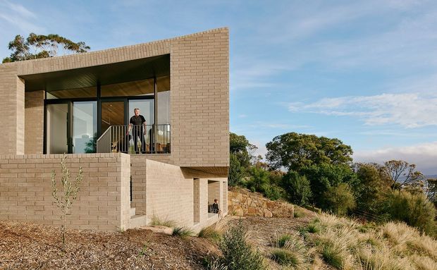 Exterior view of a contemporary two-storey residence set into a sloping, landscaped site. The house features a strong rectilinear form, pale tan brickwork, recessed balcony and entry areas, dark-framed glazing, and slender metal balcony railings. Native grasses, rockwork, and mature trees surround the building, with distant landscape visible beyond.