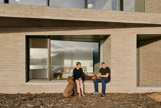 Exterior view of a contemporary multi-storey brick residence with expansive dark-framed glazing and deep projecting horizontal slab or balcony elements. Two people and a dog sit in front of a large ground-floor window, with a simple landscaped foreground. The warm pale brick facade and strong horizontal lines create a restrained, modern composition.