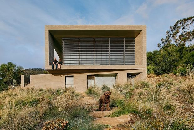 A modern two-storey rectangular house set within a landscaped coastal or bush setting. The upper level is defined by a large recessed opening enclosed with horizontal louvres, framed by a thick textured masonry or concrete surround. The lower level has solid earthy-toned walls and simple glazed openings, with a natural stone path and native grasses in the foreground.
