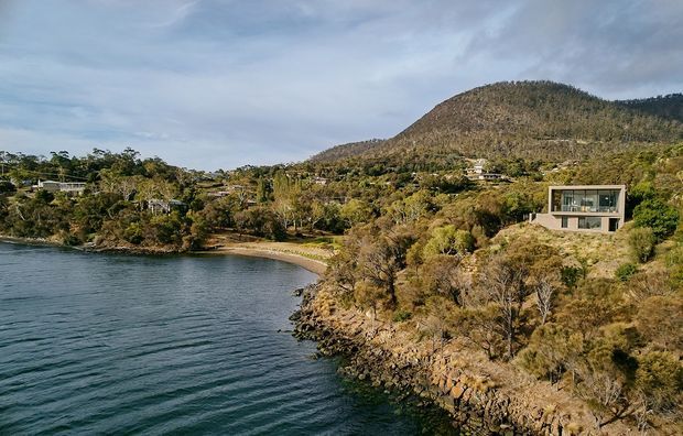 Aerial coastal landscape showing a modern rectangular house set into a wooded, rocky hillside above a small sheltered bay. The property has extensive glazing and a muted light facade, with a mountainous, forested backdrop and shoreline in the foreground.