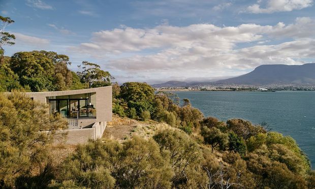 A modern concrete-and-glass house is embedded into a steep, vegetation-covered coastal or lakeside hillside. The low rectangular building has a flat roof, expansive glazing, and a projecting terrace or living space overlooking blue water and a mountainous shoreline. Native-looking scrub and trees surround the structure under a partly cloudy sky.