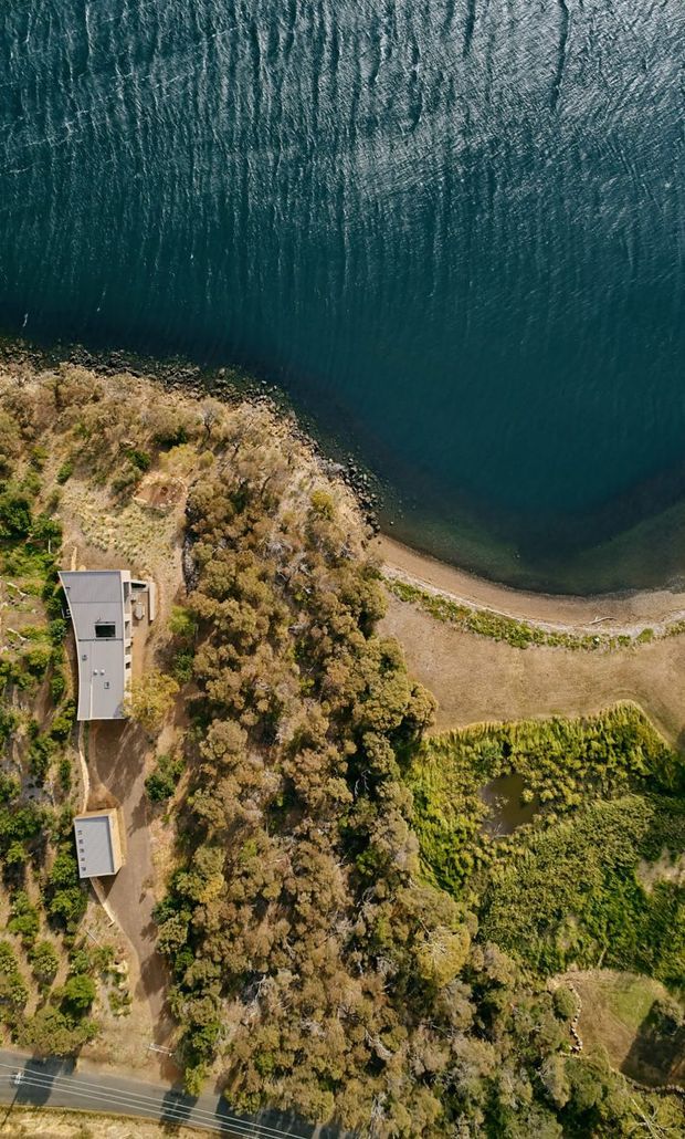 Aerial view of a property beside a large body of water, showing a narrow shoreline, wooded and dry landscaped terrain, cultivated garden areas, and two modern rectangular buildings connected by a driveway. A road runs along the lower edge of the site.