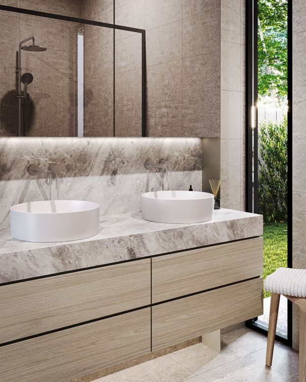 Contemporary bathroom featuring a double floating vanity with two white vessel basins, wall-mounted brushed metal faucets, and a large marble-look stone benchtop and splashback. The shower is visible behind a frameless dark metal glass screen, while full-height glazing opens toward leafy greenery. Warm timber-grain cabinetry, integrated lighting, and neutral stone surfaces create a refined, natural atmosphere.