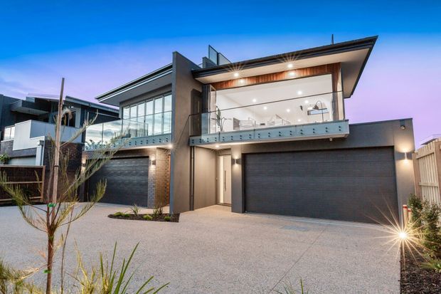 Contemporary multi-level residential facade photographed at dusk, featuring two dark garage doors, extensive upper-level glazing, a projecting flat roof, glass balustrades, timber soffit detailing, and a pale textured driveway with landscaped planting.