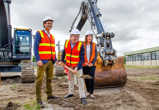 A group of three construction professionals wearing high-visibility safety vests and hard hats pose at a construction site beside a large excavator. One person holds a ceremonial shovel with soil, while heavy machinery, disturbed earth, and an industrial building are visible in the background under an overcast sky.