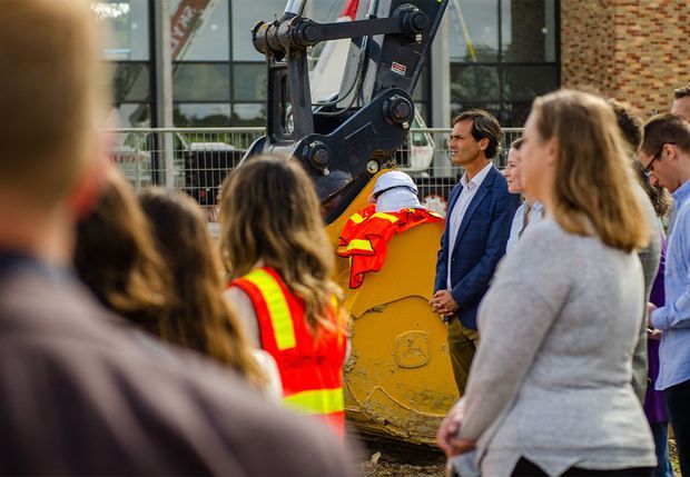 An outdoor construction-site event with a large excavator surrounded by workers and onlookers. Several people wear high-visibility safety clothing, while a man in a blue suit addresses the group. Industrial building glazing and brickwork are visible in the background.