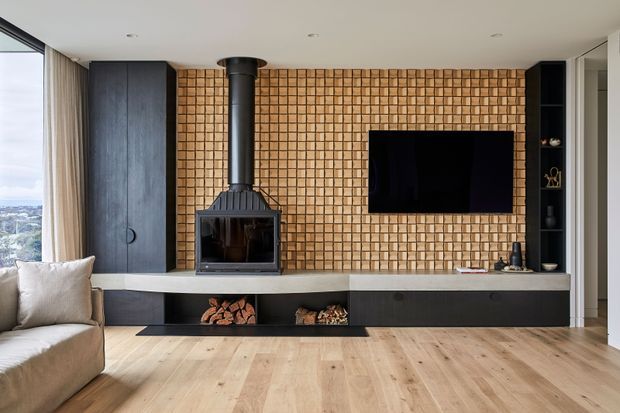 Contemporary living room centred on a black wood-burning fireplace with flue, set into a full-height textured timber feature wall. A wall-mounted television, dark built-in cabinetry, open firewood storage, pale timber flooring, and expansive glazing with city views complete the refined interior.