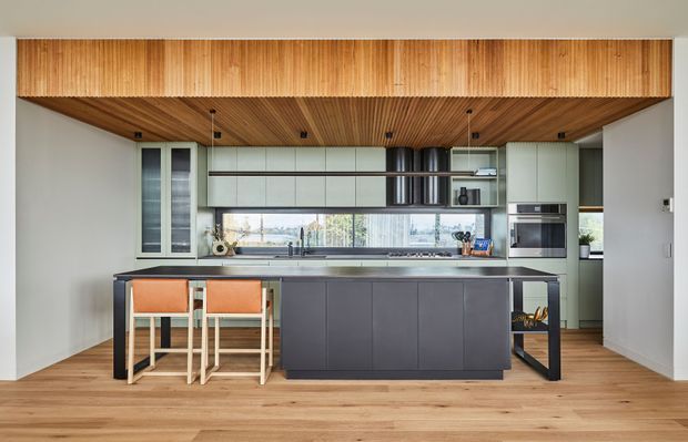 A contemporary kitchen with a large dark island and integrated seating, pale green-grey cabinetry, timber flooring, and a warm timber-lined ceiling and overhead bulkhead. Full-width glazing and a linear splashback bring in natural light, while built-in ovens and a concealed rangehood complete the streamlined layout.