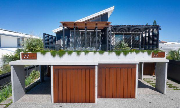 Exterior view of a contemporary multi-level residence with two timber garage doors, white rendered structural walls, a rooftop terrace or upper-level outdoor living area, timber pergola, dark vertical screening, and a sharply angled roof form. Landscaping is integrated into the terrace edge, with neighbouring homes visible in the background.