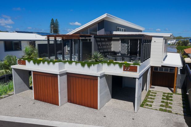Contemporary multi-level residential building viewed from the street, featuring a white rendered upper structure, dark vertical cladding, broad roof overhangs, a timber-slatted terrace pergola, rooftop planter beds, and two timber garage doors. A side driveway with grass-set concrete pavers leads to an additional garage.