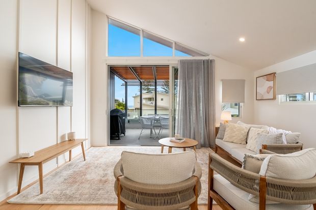 Bright contemporary living room with a large sectional sofa, woven timber-framed lounge chairs, round coffee table, timber media console and wall-mounted television. Floor-to-ceiling glazed doors open to a covered outdoor terrace, while high clerestory-style glazing and a sloping ceiling bring in abundant daylight. The room uses a soft neutral palette with light timber, textured upholstery and sheer curtains.