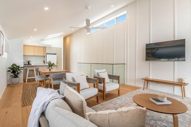 Bright open-plan living room with a neutral contemporary interior, featuring a large sectional sofa, timber-framed lounge chairs, round coffee table, wall-mounted television, timber flooring, and a compact kitchen with island visible in the background. Vertical wall panelling, clerestory glazing, recessed lighting, and a ceiling fan contribute to the architectural character.