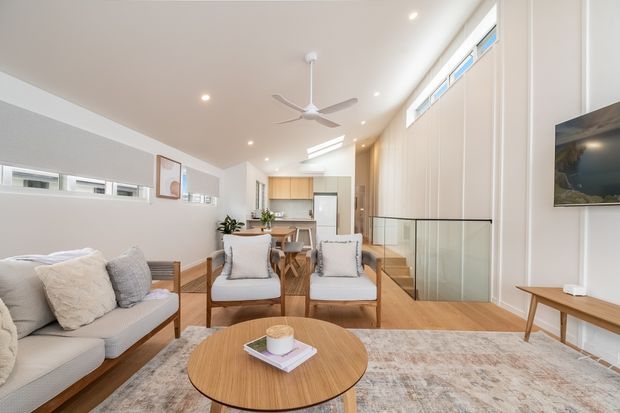 Bright open-plan living room with a neutral sofa and two timber-framed lounge chairs arranged around a round timber coffee table. The space includes light timber flooring, white walls, a high sloped ceiling with recessed lighting and a ceiling fan, clerestory-style windows, a compact dining area and kitchen in the background, and a glazed stair balustrade leading to a lower level. The interior is airy, warm, and contemporary with restrained furnishings.