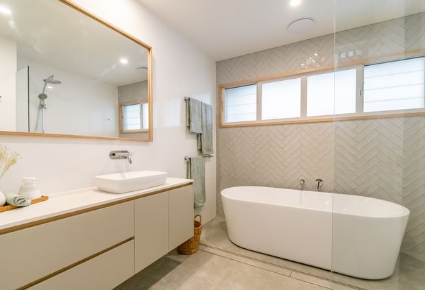 Bright contemporary bathroom with a freestanding white bathtub, wall-mounted basin vanity, large wood-framed mirror, frameless glass shower enclosure, and pale herringbone wall tile. Warm timber trim and soft neutral finishes create a clean, calm interior.