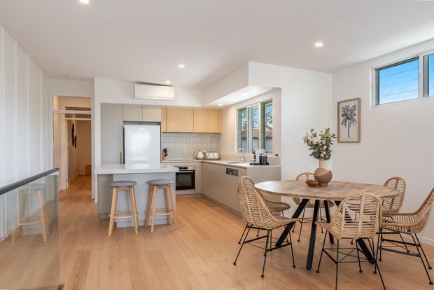 Bright open-plan kitchen and dining area with light timber flooring, white walls, pale timber cabinetry, a compact island with stools, stainless-steel appliances, and a round dining table surrounded by woven rattan-style chairs. Large windows and recessed ceiling lights create an airy contemporary coastal feel.