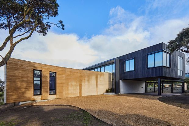 Exterior view of a contemporary two-part residence with a low timber-clad wing and a dark vertically clad upper volume projecting over a recessed ground floor. Large rectangular windows and extensive glazing connect the wings, while a broad gravel driveway and mature trees surround the house.