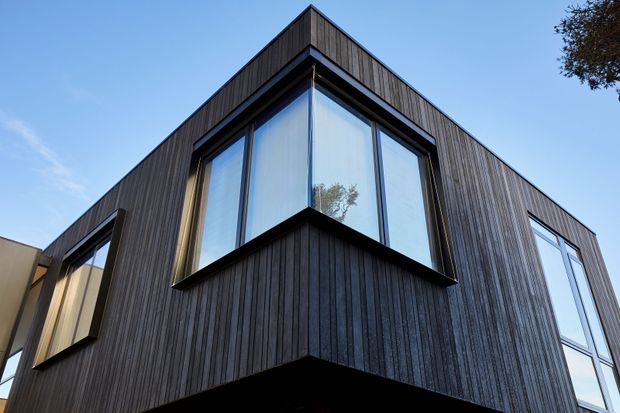 Low-angle exterior view of a contemporary multi-storey building with a sharply folded roofline and projecting corner bay windows. The facade is clad in dark vertical timber boards, with large clear glazing set in dark frames and strong geometric cantilevered forms against a blue sky.