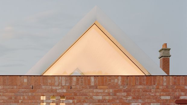 Exterior architectural view of a sharply pitched triangular gable roof rising behind a red brick boundary wall. The roof appears pale and minimalist, with warm light visible beneath the gable edge, while a traditional brick chimney stands to the right. The image has a restrained, contemporary composition with muted sky tones and warm masonry.