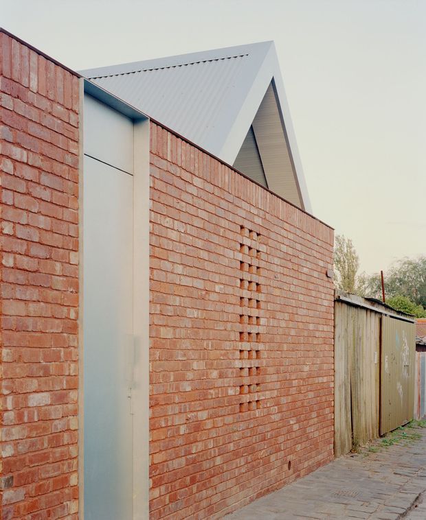 Exterior architectural view of a narrow red brick wall with a minimalist pale metal door, decorative vertical brick ventilation openings, and a sharply pitched gable roof rising behind it. The scene includes an adjacent weathered corrugated metal fence and paved walkway, with subdued neutral tones and strong geometric forms.