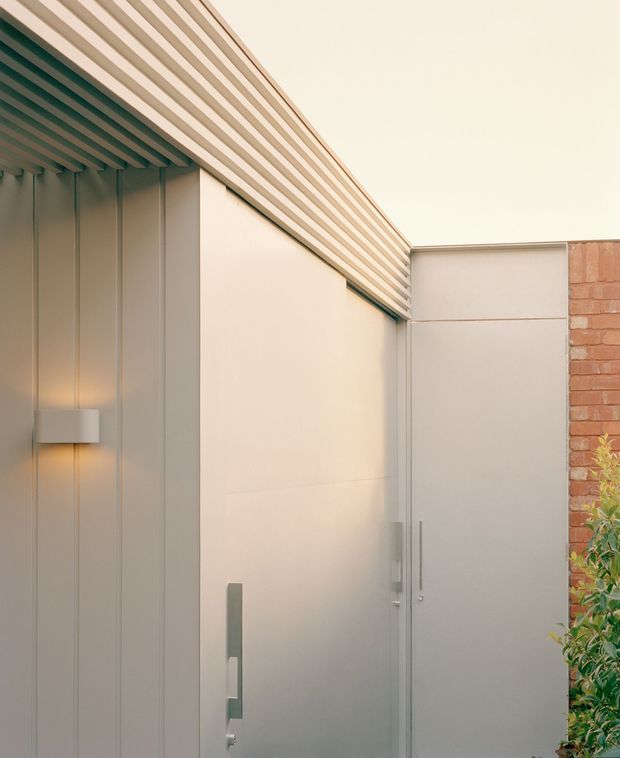 Close-up exterior architectural view of a minimalist contemporary entry or courtyard facade. Smooth pale metal wall and door panels are paired with vertical ribbed cladding, a projecting slatted roof eave, simple linear wall lighting, and a contrasting exposed red-brick boundary wall with planting at the edge.