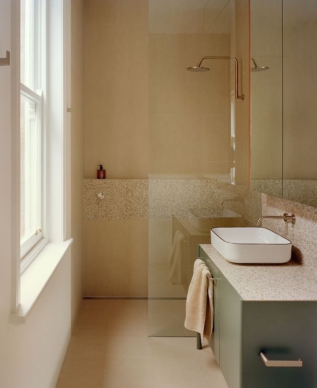 A narrow, refined contemporary bathroom with a walk-in glass shower, wall-mounted rain shower fittings, a floating green-grey vanity, white vessel basin, and warm speckled stone or terrazzo surfaces. A large white-framed window brings in natural light, while a full-height mirror visually expands the room. The palette is warm beige, cream, muted green, and brushed bronze.