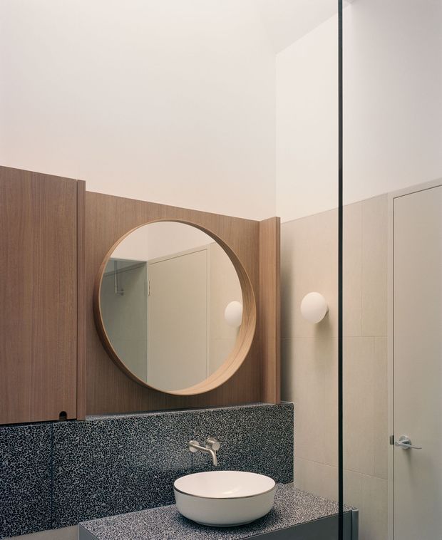 A compact contemporary bathroom with a round timber-framed wall mirror, dark speckled stone or terrazzo-style splashback and vanity surface, a white vessel basin with wall-mounted tap, pale tiled walls, and a frameless glass shower partition. Soft globe wall lighting and warm timber joinery create a refined, understated interior.
