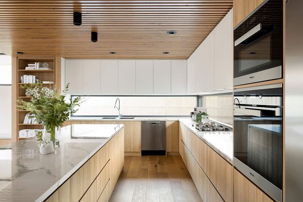 A bright contemporary galley-style kitchen with a large central island, extensive light timber cabinetry, white overhead cupboards, pale stone benchtops, integrated stainless-steel appliances, and a slatted timber ceiling. Full-height glazing and reflective surfaces enhance the spacious, clean appearance.