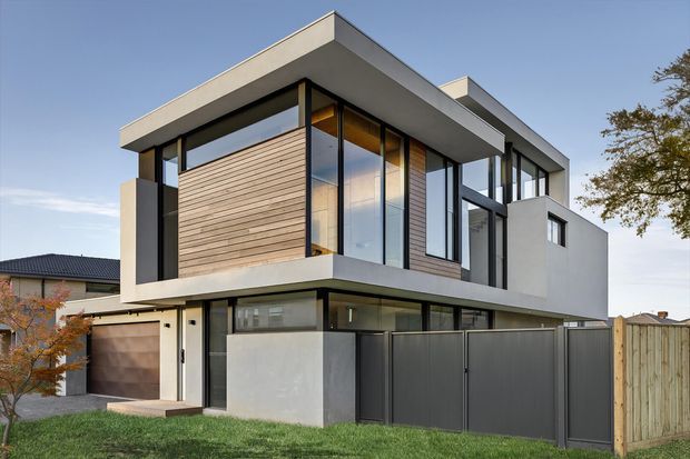 Contemporary two-storey residential house photographed from the street, featuring strong cubic forms, flat roof planes, extensive black-framed glazing, pale rendered walls, horizontal timber-look cladding, and a projecting upper-level volume. A garage and enclosed dark front boundary are visible, with neighbouring suburban homes in the background.