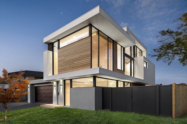 Exterior view of a contemporary two-storey home with a strong horizontal flat roof, projecting upper level, extensive black-framed glazing, white rendered walls, and a warm timber-clad feature wall. A garage, front lawn, boundary fencing, and neighbouring homes are visible.