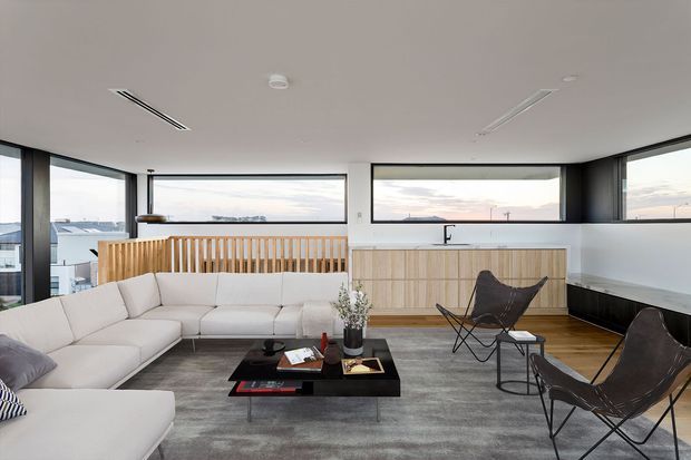 A bright, open-plan contemporary living room with expansive corner glazing and broad horizontal windows overlooking neighbouring rooftops. The room features a large pale sectional sofa, two dark leather sling chairs, a black rectangular coffee table, a grey area rug, and a built-in timber slatted screen separating the lounge from a kitchen area. The kitchen includes light timber cabinetry, a white benchtop, and a black tap. Neutral finishes, timber flooring, minimalist recessed linear vents, and abundant natural light create a refined, airy atmosphere.