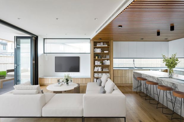Open-plan contemporary living, dining and kitchen interior with white modular seating, a round coffee table, wall-mounted television, timber built-in cabinetry and shelving, and a large marble-look kitchen island with stools. White cabinetry, timber flooring, and a slatted timber ceiling create a bright, refined space, with full-height glazing and an open door connecting to an outdoor area.