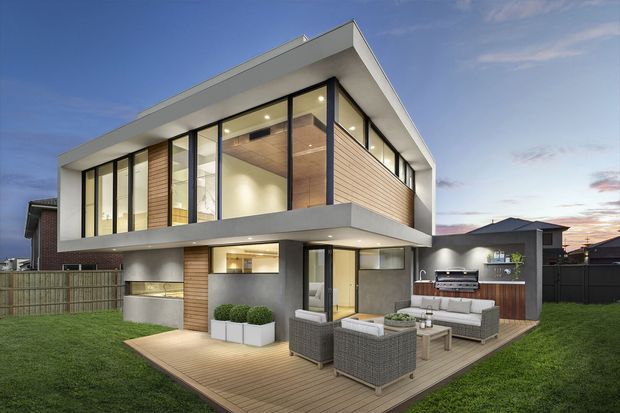 Exterior view of a contemporary two-storey home at dusk, featuring a crisp flat roof, projecting upper-level volume, extensive floor-to-ceiling glazing, dark window frames, pale horizontal timber-look cladding, and smooth light-coloured rendered surfaces. A timber deck wraps around the rear patio, furnished with an outdoor lounge setting and barbecue area. The property includes a fenced lawn and a visible neighbouring dwelling.