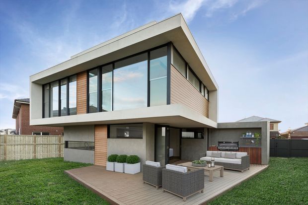 Contemporary two-storey residential house viewed from the rear, featuring a projecting upper level with broad floor-to-ceiling glazing, pale horizontal roof slab, dark window frames, and warm vertical timber-look wall cladding. The lower level opens onto a timber deck with outdoor lounge furniture, planters, and a built-in barbecue area. A fenced suburban yard surrounds the home.