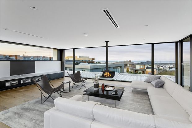 A bright, contemporary open-plan living room with expansive corner glazing overlooking a marina and surrounding buildings. The room features a large white sectional sofa, two black leather sling chairs, a low black coffee table, a wall-mounted television with built-in cabinetry, pale timber flooring, and a suspended central fireplace with a flue. The space has a clean, refined, minimalist look and a standard-to-high ceiling with generous glazing.