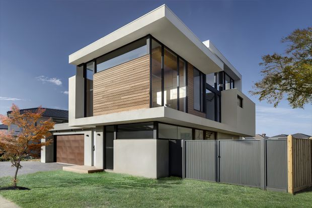 A contemporary two-storey residential house photographed from the street, featuring a strong cantilevered upper level, crisp white rendered surfaces, extensive dark-framed glazing, and a horizontal light timber-clad feature wall. The ground level includes a garage, entry, privacy walls, and a tall metal gate and fence. The home has a flat roof and clean geometric massing, set within a suburban landscape.
