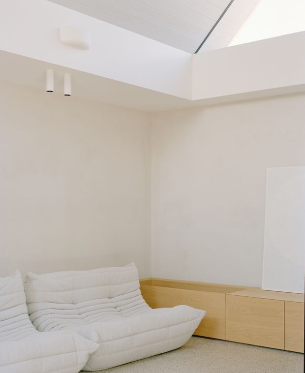 Minimalist, light-filled living room with a low white modular sofa, pale beige walls, light timber built-in cabinetry, and a white slatted vaulted ceiling with clerestory-style glazing. The space has a clean, understated contemporary character and a soft neutral palette.