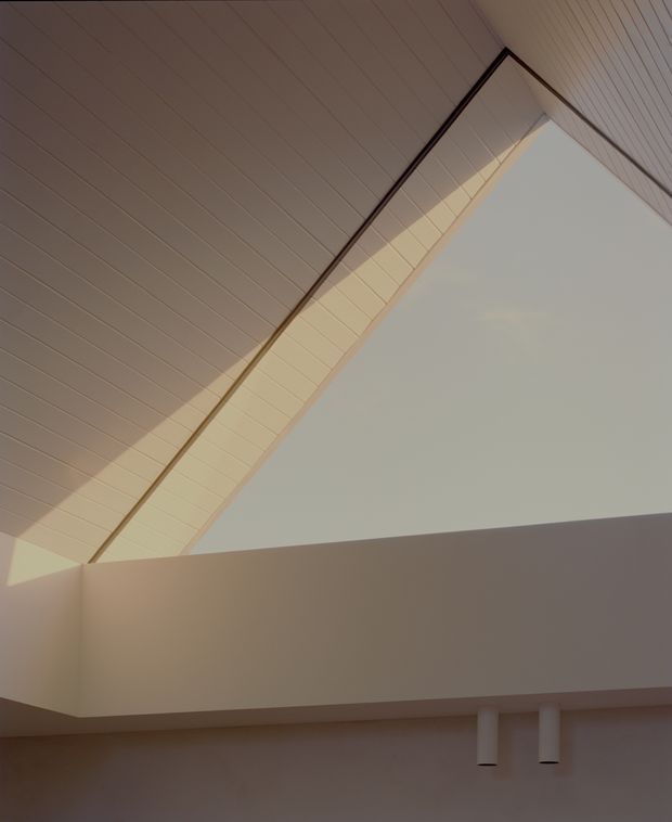 Interior architectural detail showing a dramatic high sloped ceiling with pale timber or painted linear paneling, a large triangular clerestory or skylight opening, and a deep white architectural bulkhead casting soft shadows. Minimal cylindrical wall-mounted lights are visible below. The composition emphasizes natural light, angular geometry, and a calm neutral palette.