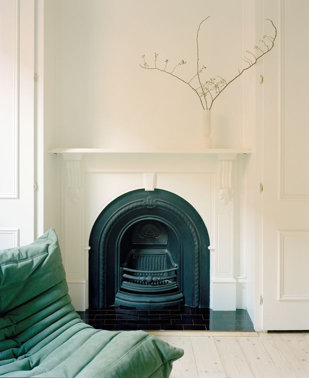 A refined interior vignette centered on an ornate black cast-iron fireplace with a decorative white mantel and panelled surround. Pale cream walls and detailed mouldings create a traditional architectural backdrop, while a sage-green quilted lounge chair occupies the foreground. A simple vase with delicate branches sits on the mantel, and pale timber floorboards complete the calm, elegant composition.