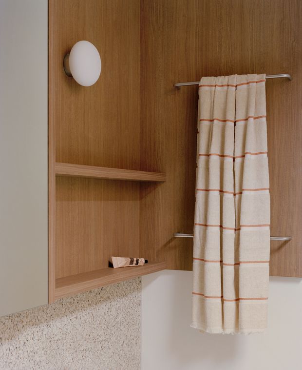 Close-up view of a warm-toned bathroom featuring vertical timber-look wall panelling and open built-in shelving, a white wall sconce, chrome towel rails with a cream-and-rust checked towel, and a terrazzo-style wall surface below. The composition focuses on built-in bathroom storage and finishes.