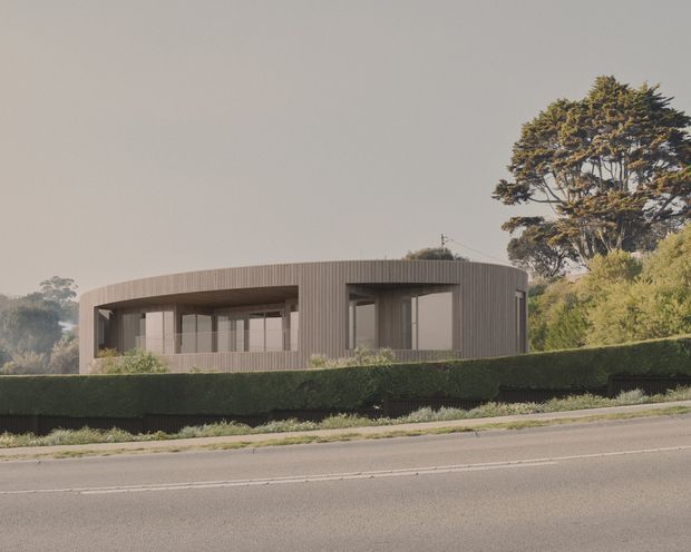 A low-profile contemporary house viewed from across a roadway, featuring a rounded rectangular form, continuous dark vertical cladding, extensive floor-to-ceiling glazing, and a flat or very gently curved roofline. The home is screened by neatly clipped hedges and mature trees, creating a restrained suburban streetscape.