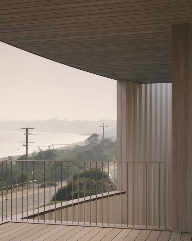 A minimalist covered balcony or terrace framed by warm vertical timber-clad walls and a dark timber-lined soffit. Slim metal balustrades overlook a sloping suburban or coastal landscape with utility poles and distant water or haze. The restrained palette and strong horizontal and vertical lines create a calm, contemporary architectural composition.