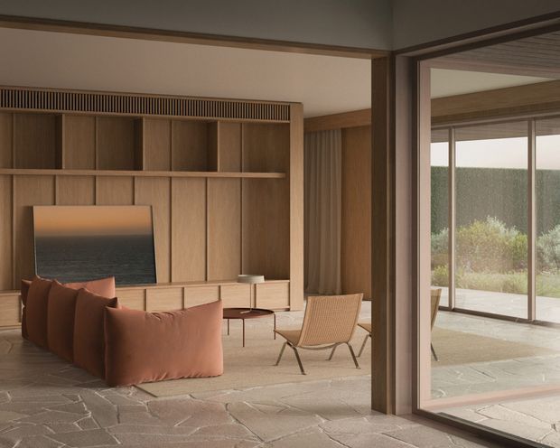 Warm, refined contemporary living room with full-height timber built-in shelving and media cabinetry, a large low rust-coloured sofa, woven lounge chair, slim coffee tables, and a textured stone or tile floor. Floor-to-ceiling glazing opens to a paved outdoor terrace and landscaped garden. The room uses strong indoor-outdoor connections, natural timber, earthy upholstery, and restrained detailing.