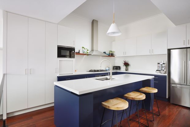 Bright, spacious contemporary kitchen with a large white island featuring deep navy-blue cabinetry and an integrated sink. White floor-to-ceiling cabinetry surrounds built-in appliances, with stainless-steel refrigeration, a stainless rangehood, open shelving, and a white pendant light. The room has polished warm timber flooring and a clean, high-contrast palette, with timber stools arranged at the island.
