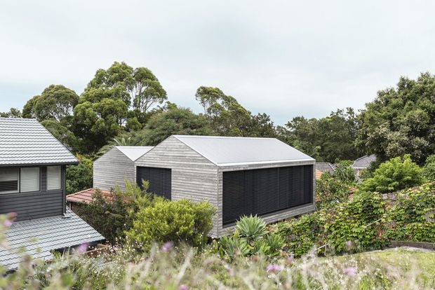 Exterior architectural photograph of a compact contemporary building set within a lush, landscaped residential setting. The structure has a simple rectangular form, a low-pitched gable roof, horizontal light-grey weatherboard cladding, and large dark louvred or screened openings. Other dark-clad pitched-roof buildings are partially visible among mature trees and garden planting.