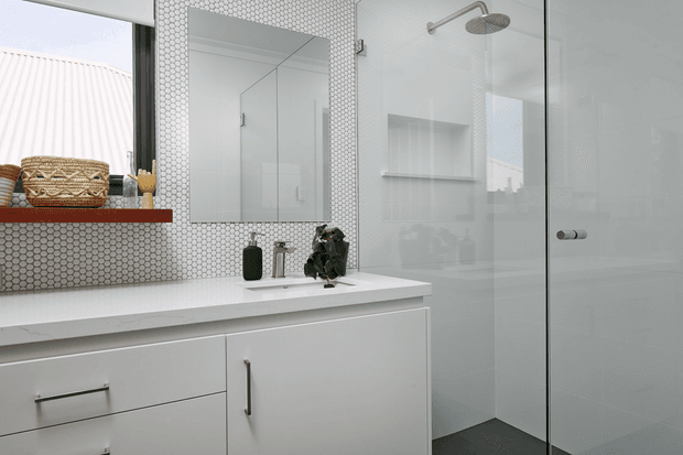 Bright contemporary bathroom with a white floating vanity, integrated white benchtop, large mirror, and walk-in glass shower. The vanity wall is finished in small white hexagonal mosaic tiles, while the shower has pale large-format wall tiles and a dark floor. A timber shelf with woven baskets adds warmth and texture.
