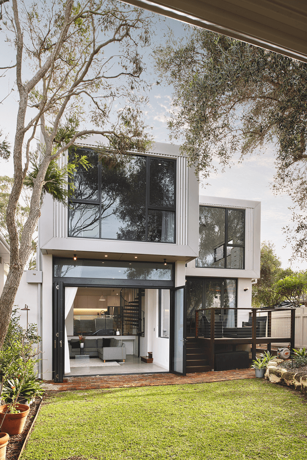 A contemporary two-storey residential extension viewed from a landscaped backyard. The white-clad façade features large black-framed windows, expansive glazed openings, an upper-level projecting volume, and a covered outdoor deck or terrace with dark metal balustrading. Through the open ground-floor glazing, a modern living area and kitchen are visible, including a spiral staircase. Mature trees, lawn, garden beds, and brick paving surround the house.