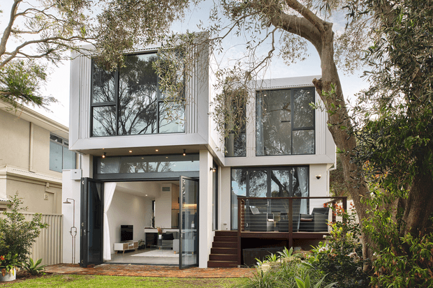 A contemporary multi-level residential extension viewed from the landscaped front yard. The white rendered facade features strong vertical ribbed detailing, large black-framed glazed openings, projecting upper volumes, and a broad open connection to the ground-floor living area. Dark timber steps and decking lead through a leafy garden setting, with mature trees partially framing the house.