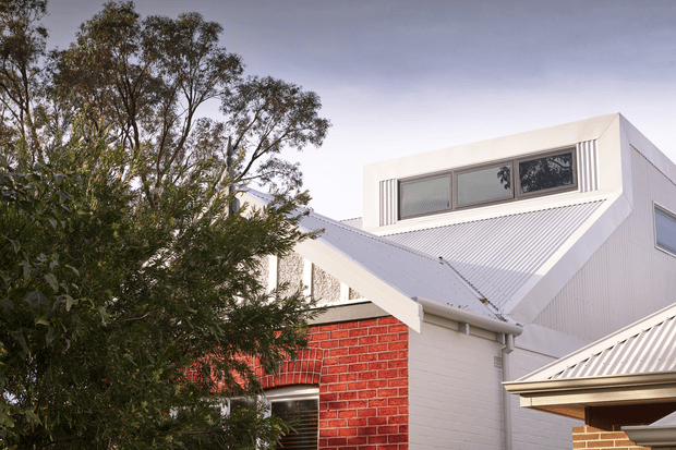 Exterior architectural view of a contemporary multi-level residence, featuring white corrugated metal roofing and cladding, a prominent red brick wall, projecting clerestory-style windows, and layered pitched roof forms. Mature trees partially screen the facade.