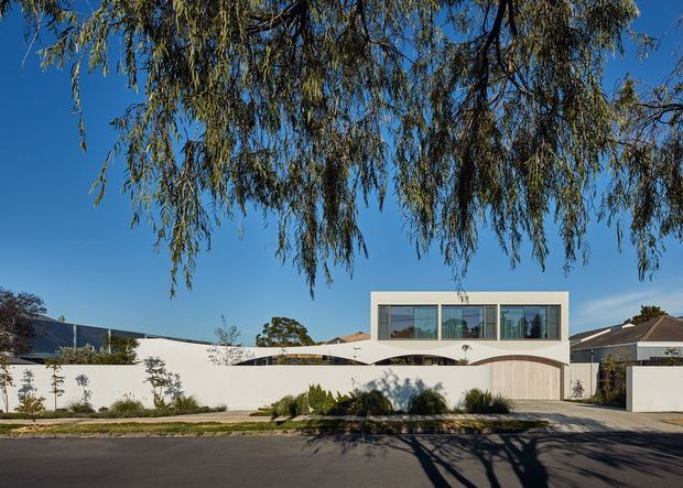 Street-facing view of a contemporary two-storey residence with a pale rendered facade, curved white boundary wall, large upper-level rectangular windows, and a timber-toned garage door. Low landscaped planting sits behind the street verge, while a mature overhanging tree frames the scene against a clear blue sky.